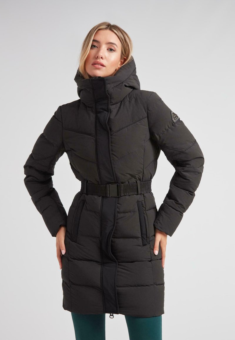 Kvinde iført en lang sort quilted frakke med høj krave og bælte, stående med hænderne på hoften mod en ensfarvet lys baggrund.