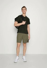 Tricou texturat de culoare verde închis, pantaloni scurți olive cu logo negru, pantofi sport albi și un rucsac negru purtat pe un umăr.
