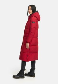 Rød lang pufferjakke med hætte, prydet med vandret quiltede sektioner, sidelommer og et patch-logo på venstre arm. Båret med sorte støvler.