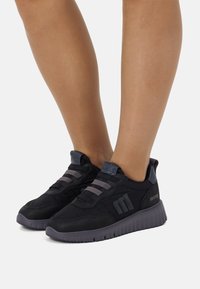 Zapatillas negras con parte superior texturizada en negro, detalles en gris y cordones elásticos. Presentan un collar acolchado y una suela gruesa de goma gris.