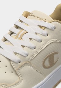 Μπεζ και λευκό αθλητικό παπούτσι με λευκά κορδόνια, μπεζ λογότυπο Champion στο πλάι και το όνομα της μάρκας Champion κεντημένο στη γλώσσα.