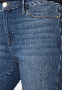 Tummansiniset denimfarkut, joissa on sileä pinta, etutaskut, kullanväriset ompelukoristeet ja nahkainen merkkilappu vyötäröllä.