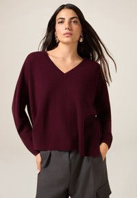 Maglione bordeaux con scollo a V in morbido tessuto lavorato a maglia con maniche larghe, abbinato a pantaloni grigi con tasche laterali e vestibilità comoda.
