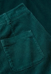 Tela de pana verde oscuro con acanaladuras verticales, que presenta un bolsillo trasero con costuras reforzadas, mostrando textura y una rica profundidad de color.