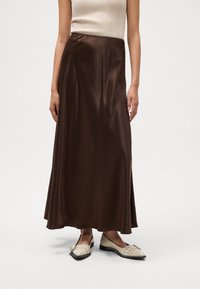 Jupe maxi en satin brun avec une texture lisse et un léger éclat, associée à des chaussures plates de couleur crème avec des accents décoratifs.