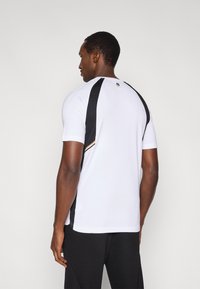 Λευκό αθλητικό πουκάμισο με μαύρες πλαϊνές επιφάνειες και χρυσή λεπτομέρεια, κοντομάνικο, με λείο ύφασμα, στρογγυλή λαιμόκοψη, απεικονίζεται από πίσω.