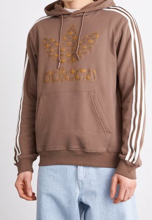 Homme portant un sweat à capuche Adidas marron avec un logo doré et des bandes blanches sur les manches, associé à un jean bleu clair.