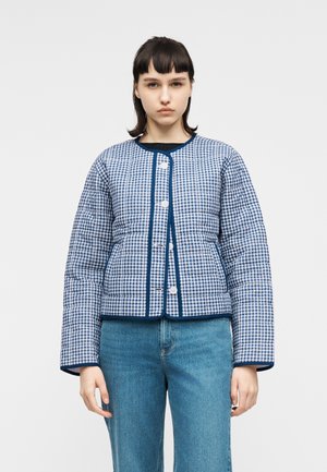 Jeune femme aux cheveux courts et foncés portant une veste matelassée à carreaux bleus et blancs ainsi qu'un jean bleu clair, debout devant un fond uni.