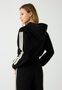 Felpa nera con cappuccio e strisce crema sulle maniche, texture morbida, design aderente, tasche frontali, vita cintrata per un look sartoriale.