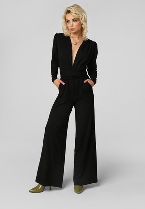 Blonde Frau trägt einen schwarzen langärmeligen Overall mit tiefem V-Ausschnitt, weiter Beinform und grünen spitzen High-Heels, Hände in den Taschen.