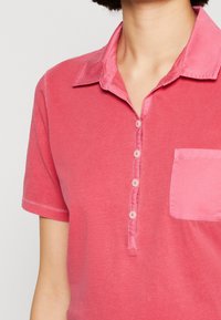 Růžová polo košile s krátkým rukávem, s knoflíkovou légou a náprsní kapsou v mírně světlejším odstínu růžové.