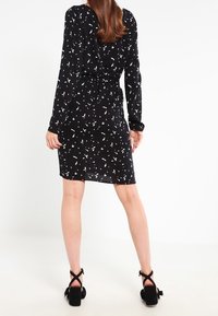 Robe noire à taille cintrée, manches longues et ourlet atteignant le genou. Présente un motif abstrait blanc dispersé. Portée avec des talons noirs à brides.