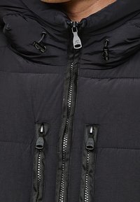 Gros plan d'une veste noire rembourrée avec trois fermetures éclair verticales et des cordons réglables près du col.