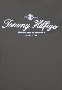 Tissu gris avec un texte blanc affichant "Tommy Hilfiger," "EST. 1985," "MARQUE DÉPOSÉE," et "NEW YORK." Texte dans une police cursive.