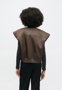 Καφέ αμάνικο μπουφάν με διαμορφωμένο σχέδιο, λεία υφή και εφαρμοστό τελείωμα. Ορατές ραφές στο πίσω μέρος.