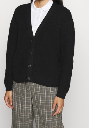 Kobieta ubrana w czarny sweter na guziki, pod którym ma białą koszulę z kołnierzykiem, oraz szare spodnie w kratę, stojąca na jednolitym tle.