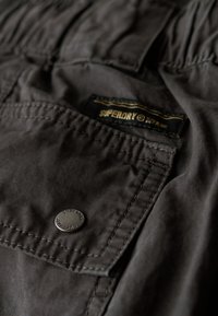 Dunkelgrauer Stoff mit sichtbaren Nähten und einer Fronttasche. Verfügt über einen Metallknopf und ein angenähtes Markenetikett mit schwarzem und goldenem Text.