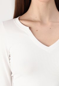 Bijela rebrasta bluza dugih rukava s V-izrezom, s malo skraćenim krojem i teksturiranim materijalom.