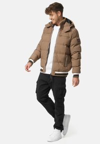 Brown puffer jacket with hood, zip closure, two front pockets, contrasting striped cuffs and hem, paired with black cargo pants and white sneakers.
