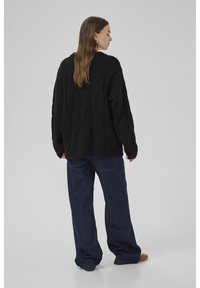 Frau steht seitlich und trägt einen schwarzen Strickpulli, dunkle weiter geschnittene Jeans und braune Schuhe vor einem grauen Hintergrund.