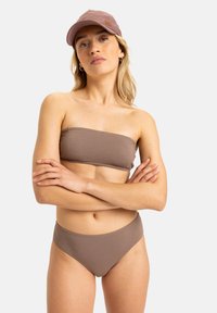 Brassfreier brauner Bikini aus strukturiertem Stoff, kombiniert mit einer passenden braunen Kappe. Der Slip hat einen mittelhohen Schnitt. Das Model posiert mit verschränkten Armen.