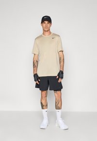 Homme portant un shirt beige Nike, un short noir, une casquette noire, des gants sans doigts, des chaussettes blanches Nike et des baskets blanches, se tenant devant un fond uni.