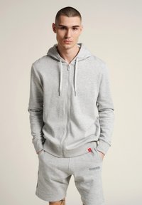 Helles graues Zip-Hoodie mit einer Fronttasche, verstellbarem Kordelzug und gerippten Bündchen. Kombiniert mit passenden Shorts mit Logo-Details.
