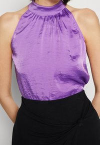 Top halter in satin viola con collo alto, caratterizzato da dettagli arricciati sullo scollo e una texture liscia e brillante. Gonna nera annodata in vita.