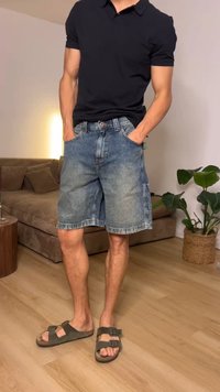 Άντρας με μαύρο πόλο μπλουζάκι, τζιν σορτς και καφέ σανδάλια, στέκεται με τα χέρια στις τσέπες σε ένα καθιστικό.