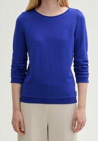 Niebieska, teksturowana koszulka z długim rękawem, dekoltem typu crew i prążkowanymi mankietami oraz dołem. Posiada subtelny wzór oraz detale z przeszyciami bocznymi.