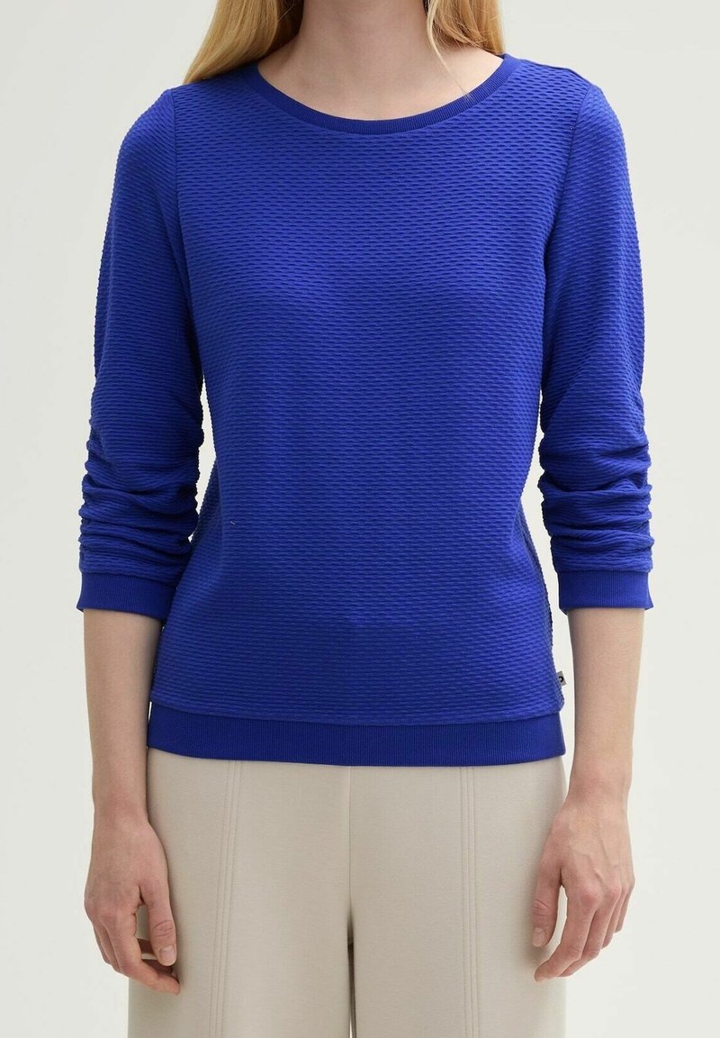 Niebieska, teksturowana koszulka z długim rękawem, dekoltem typu crew i prążkowanymi mankietami oraz dołem. Posiada subtelny wzór oraz detale z przeszyciami bocznymi.