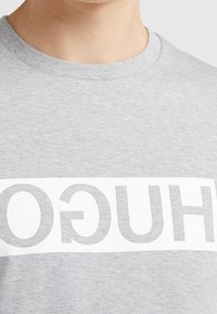 Grauer Pullover mit Rundhalsausschnitt, der ein weißes horizontales Band mit der Marke "HUGO" in fetten grauen Buchstaben zeigt. Baumwoll-Mischgewebe.