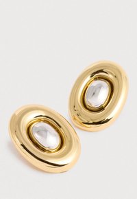 Kuldseid ovaalseid kõrvarõngaid, millel on siledalt tõstetud keskus läikivas hõbedas, asetatuna lihtsale valgele pinnale.