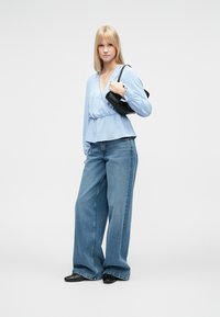 Lyseblå slå-om-bluse, høj talje med rynkedetalje, kombineret med vide denimjeans. Sort håndtaske og flade sko fuldender looket.