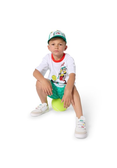 Garçon portant une casquette de baseball, un t-shirt blanc avec un dessin animé, un short vert et des baskets blanches, assis sur une grosse balle de tennis jaune sur fond blanc.