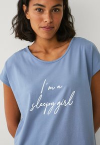 Lyseblå t-shirt med korte ærmer, der har en rund halsudskæring og hvid tekst med skrifttype, der står "Jeg er en søvnig pige" på forsiden.