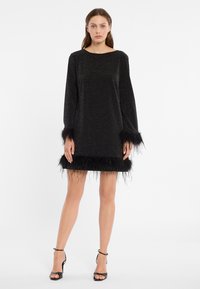 Zwarte jurk met lange mouwen, met een glinsterende textuur en een veerafwerking bij de manchetten en de zoom. Geaccentueerd met zwarte hoge hakken.