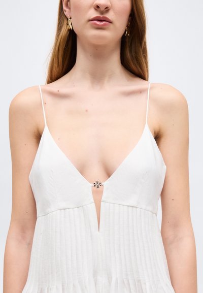 Žena v bielych šatách bez rukávov s tenkými ramienkami a hlbokým výstrihom do V, ktorý drží malá kovová pracka.