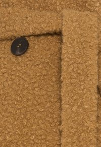 Brązowa, teksturowana tkanina o splocie boucle. Widoczny jest czarny guzik, który dodaje detalu materiałowi, który wydaje się miękki i ciepły.