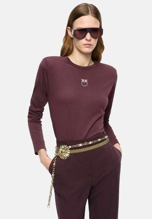 Donna che indossa un top a maniche lunghe viola scuro e pantaloni, occhiali da sole oversize e una cintura a catena dorata con un grande dettaglio floreale, mani in tasca.