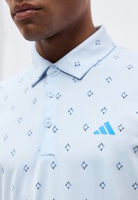 Κοντινό πλάνο ενός ατόμου που φορά ένα γαλάζιο πουκάμισο με γιακά, διακοσμημένο με μικρά σκούρα μπλε σχέδια σε σχήμα σταυρού και ένα γαλάζιο τρίγωνο λογότυπο στο στήθος.