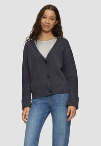 Dunkelgrauer Cardigan mit tiefem V-Ausschnitt, gerippter Textur und vier Knöpfen. Mit weiten Ärmeln und lockerer Passform, kombiniert mit hellblauen Jeans.
