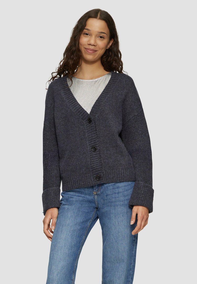 Dunkelgrauer Cardigan mit tiefem V-Ausschnitt, gerippter Textur und vier Knöpfen. Mit weiten Ärmeln und lockerer Passform, kombiniert mit hellblauen Jeans.