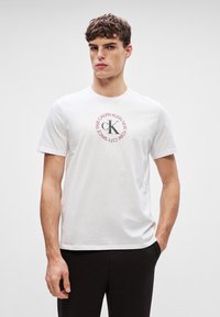 T-shirt de algodão branco com uma gráfica circular com "CALVIN KLEIN NEW YORK CITY SINCE 1988" e um logótipo central "ck" em cinza. Mangas curtas.