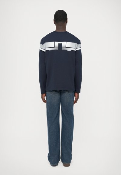 Μπλε σκούρο πουκάμισο με μακριά μανίκια και λευκή ριγέ σχεδίαση στην πλάτη, σε συνδυασμό με άνετο μπλε τζιν και καφέ παπούτσια.