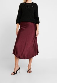 Pull noir avec accents perlés, associé à une jupe midi en satin bordeaux avec un ourlet fluide et une fente latérale, complété par des talons noirs à brides.