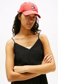 Rote Baseballmütze mit einem Logo-Patch, kombiniert mit einem schwarzen Trägertop mit feiner Spitzenborte und dünnen Trägern. Glatte Stofftextur.