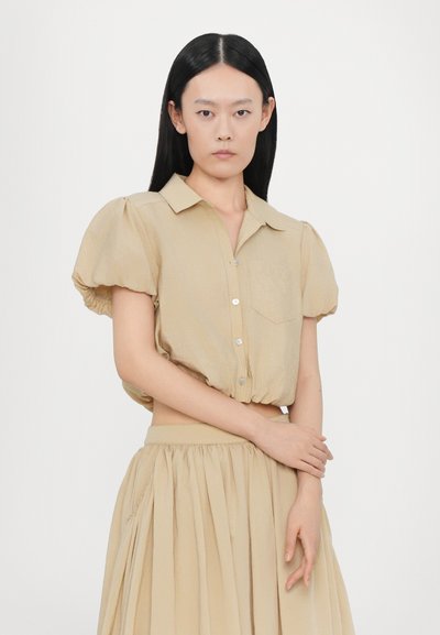 Beige, kurz geschnittene Bluse mit Knöpfen, Puffärmeln, Brusttasche und weicher Textur; kombiniert mit einem passenden, beige fließenden Rock.