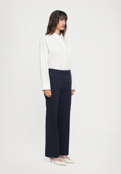 Chemise blanche à manches longues, associée à un pantalon large bleu marine. Le modèle porte des escarpins crème à bout pointu. Design simple, texture lisse.