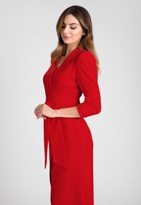Czerwona dopasowana sukienka z wykładanym kołnierzykiem, rękawami o długości trzech ćwiartek i zawiązywanym paskiem w talii. Gładki materiał i detal w postaci bocznego rozcięcia podkreślają design.
